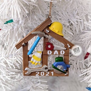May include: A brown and tan Christmas ornament shaped like a house, featuring tools and the word "DAD". Includes a yellow hard hat, blue hammer, and the year "2021". The ornament is hanging on a white Christmas tree.