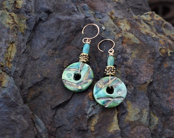 May include: Handmade earrings featuring turquoise and green ceramic discs with etched designs. The earrings have gold-colored ear wires, turquoise beads, and decorative gold-colored metal beads. The earrings are displayed on a dark, textured surface.