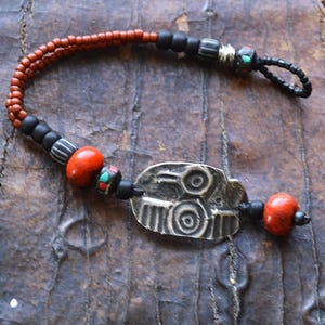 Op de afbeelding: Handgemaakte armband met een zilverkleurige metalen hanger met cirkelvormige ontwerpen, geaccentueerd door rode, zwarte en patroon kralen. De armband heeft een lus sluiting en wordt gepresenteerd op een getextureerd bruin oppervlak.