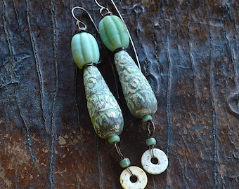 May include: A pair of handcrafted earrings featuring a combination of green and cream-colored beads. The earrings have a long, dangling design with intricate patterns and textures, suspended from silver-tone ear wires.