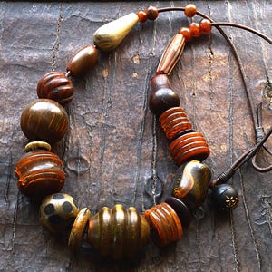 Può includere: Collana artigianale con perline di legno nei toni del marrone, ambra e crema. Le perline hanno forme e dimensioni diverse, tra cui design rotondi, a barile e allungati. La collana è infilata su un cordino marrone.