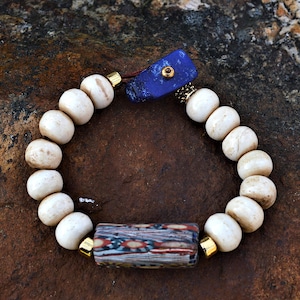 以下が含まれることがあります： クリーム色の丸いビーズ、長方形の模様のビーズ、青い石のアクセントが特徴の手作りのブレスレット。ブレスレットは茶色のコードに通されており、金色の金属アクセントが付いています。