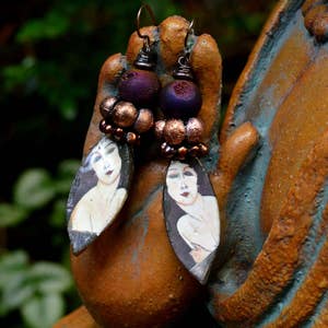 May include: Art Deco earrings with oval pendants depicting painted portraits of women. The earrings have purple and bronze beads and copper-coloured ear wires. The background is a blurred green and brown.