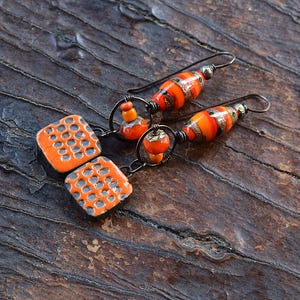 May include: Handmade orange and black earrings. The earrings feature square, textured orange ceramic beads and elongated orange and gold striped glass beads. The earrings are connected with dark metal wire and hooks.