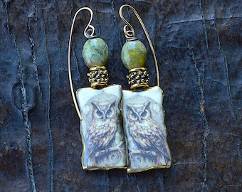 May include: A pair of handcrafted earrings featuring an owl design. Each earring has a rectangular pendant with an owl illustration, a green bead, and gold-colored accents. The earrings hang from gold-colored ear wires.
