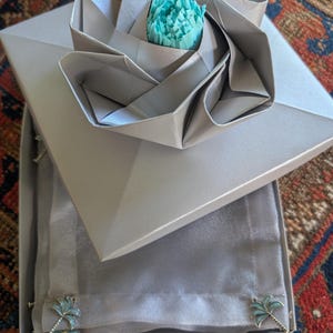 May include: A square silver gift box topped with a folded paper rose featuring a bright turquoise center. Inside the box, a folded silver fabric item is visible, adorned with two small silver and turquoise palm tree-shaped metal embellishments.