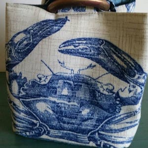 May include: A white canvas tote bag with a blue and white crab design. The bag has a brown fabric handle with a wooden ring.