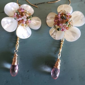 May include: A pair of flower-shaped earrings. Each earring features a white, petal-like design with a cluster of colorful beads at the center. A gold wire connects the flower to a pink, teardrop-shaped gem. The earrings are set against a teal background.