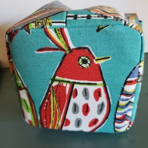 May include: A teal blue fabric tote bag featuring a colourful bird design. The bird is red, yellow and white with black eyes and a long beak. The bag has a zipper closure and a handle.