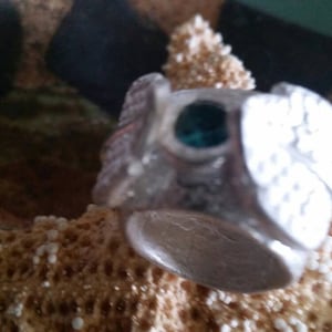 May include: A silver ring with a green stone set in the center. The ring is decorated with intricate designs and has a wide band.
