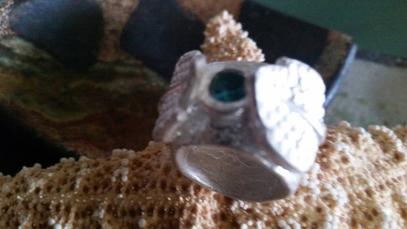 May include: A silver ring with a green stone set in the center. The ring is decorated with intricate designs and has a wide band.