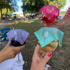 May include: Three wine glasses with different colored wine, each covered with a sheer fabric wine glass cover with a decorative charm. The covers are lavender, turquoise, and pink.