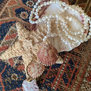 May include: A white pearl necklace with a gold pendant featuring three layered seashells. The necklace is displayed on a seashell and starfish.