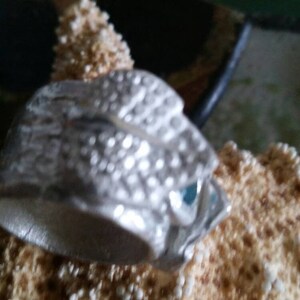 May include: A silver ring with a detailed textured design resembling scales. The ring has a blue gemstone set into the design.