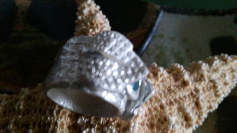 May include: A silver ring with a detailed textured design resembling scales. The ring has a blue gemstone set into the design.