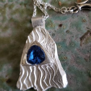 May include: A silver pendant necklace with a blue gemstone set in a triangular shape. The pendant has a textured surface with wavy lines.