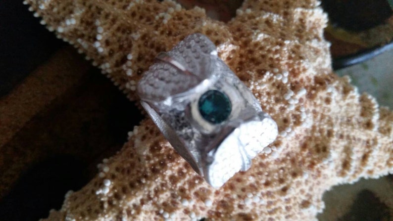May include: A silver ring with a teal gemstone set in the center. The ring is decorated with a detailed design of leaves and swirls.