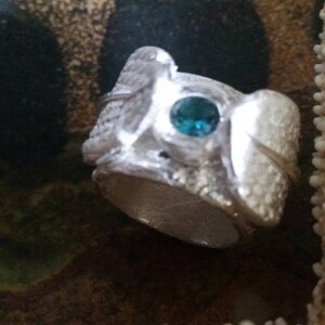 May include: A silver ring with a textured surface and a teal blue gemstone set in the center.