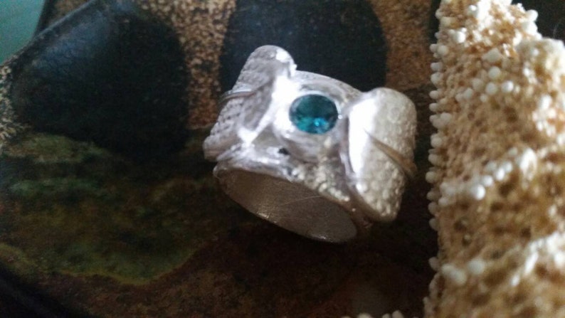 May include: A silver ring with a textured surface and a teal blue gemstone set in the center.
