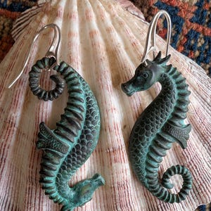 May include: A pair of seahorse earrings with a verdigris finish, featuring intricate detailing. The earrings are displayed on a large seashell, showcasing their aquatic theme. The hooks are silver-toned.