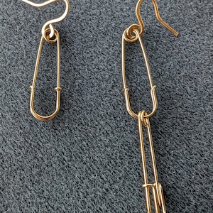 May include: A pair of gold safety pin earrings. The earrings are made of two safety pins that are connected together. The safety pins are about 2 inches long.