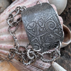 May include: A silver-toned rectangular pendant necklace featuring embossed seashells and starfish. The pendant is connected to a twisted chain necklace with a lobster clasp. The jewelry is displayed on a seashell.