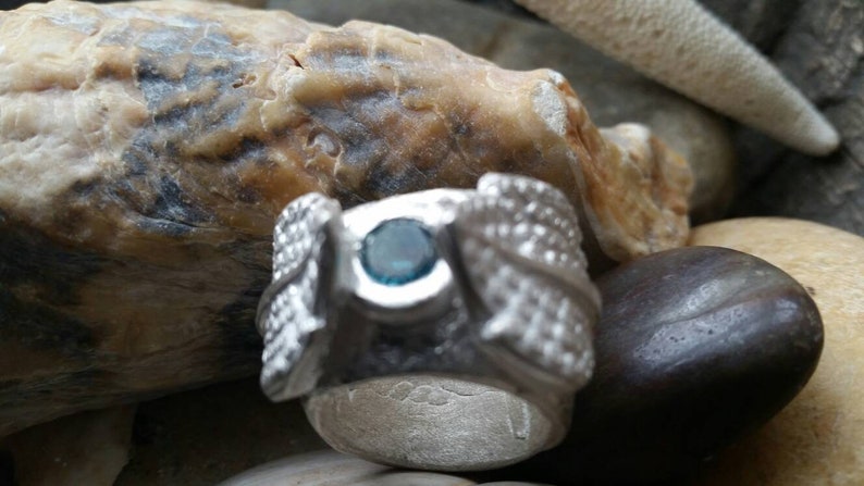 May include: A silver ring with a blue gemstone set in the center. The ring is decorated with intricate designs on the sides.