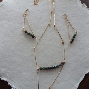 May include: A gold necklace and earrings set with green and black gemstone beads. The necklace has a long, delicate chain with a geometric design. The earrings are small and triangular.