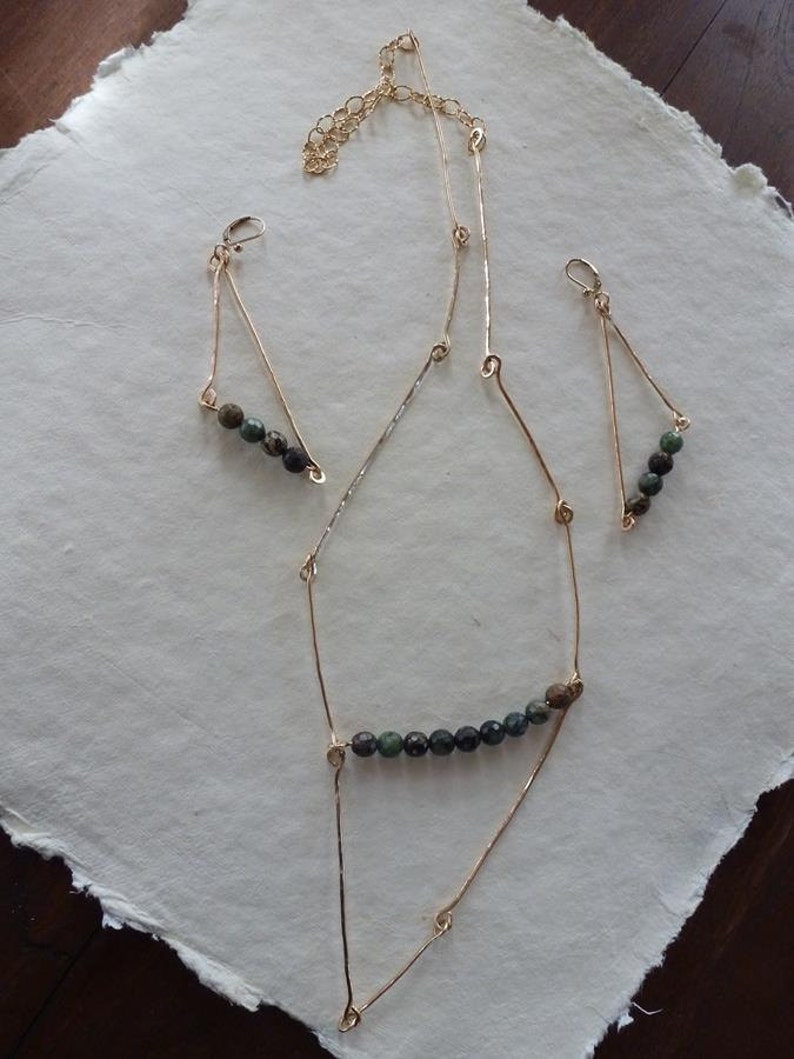 May include: A gold necklace and earrings set with green and black gemstone beads. The necklace has a long, delicate chain with a geometric design. The earrings are small and triangular.