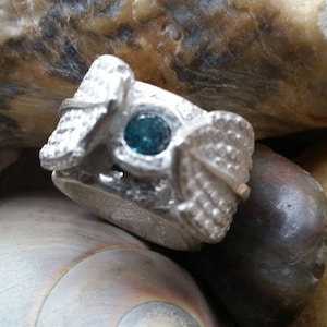 May include: A silver ring with a textured design featuring a blue gemstone. The ring is set against a background of rocks.