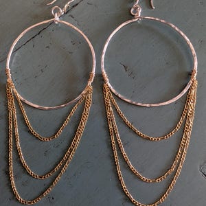 May include: A pair of silver hoop earrings with three strands of gold chain dangling from the bottom. The earrings have a hammered texture on the silver hoops and are displayed against a teal background.