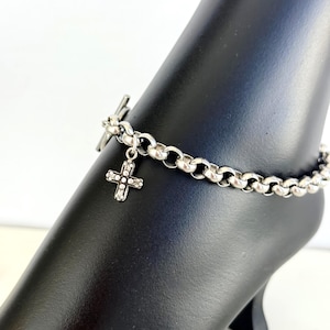 May include: Silver-toned anklet featuring a cross pendant. The anklet has a chain-link design with a toggle clasp. The cross pendant is adorned with small, sparkling embellishments. The anklet is displayed on a black mannequin foot.