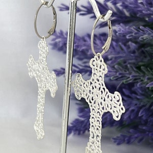 May include: Silver filigree cross earrings with a delicate, intricate design. The earrings are hanging from a silver hoop.