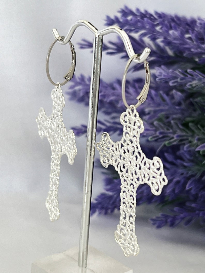 May include: Silver filigree cross earrings with a delicate, intricate design. The earrings are hanging from a silver hoop.