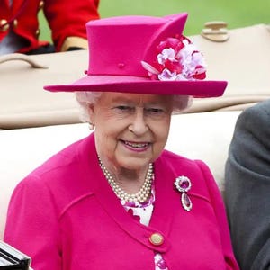 May include: A woman wearing a pink suit and a pink hat with a flower embellishment. She is smiling and has a pearl necklace and a jeweled brooch.