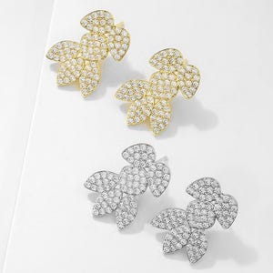 May include: Two pairs of leaf-shaped stud earrings. One pair is gold-toned, and the other is silver-toned. Each earring features three leaves encrusted with small, clear crystals, creating a sparkling effect. The earrings are displayed on a white surface.