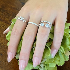 May include: A hand displaying four silver rings. The rings include a delicate "love" script ring, a simple band, and a double-stone ring with a beaded band. The hand rests on a bed of green leaves.