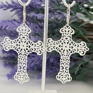 May include: Silver filigree cross earrings with a delicate, intricate design. The earrings are hanging from a silver stand.