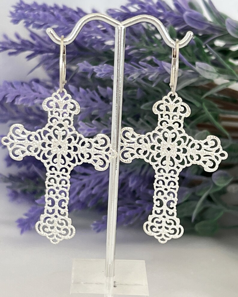 May include: Silver filigree cross earrings with a delicate, intricate design. The earrings are hanging from a silver stand.