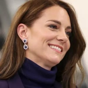 May include: A woman with long brown hair wearing a dark blue turtleneck sweater and a pair of dangling earrings with blue gemstones.
