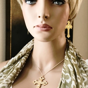 May include: A gold necklace with a large, ornate cross pendant. The necklace is worn by a person with blonde hair and a green and white patterned scarf.
