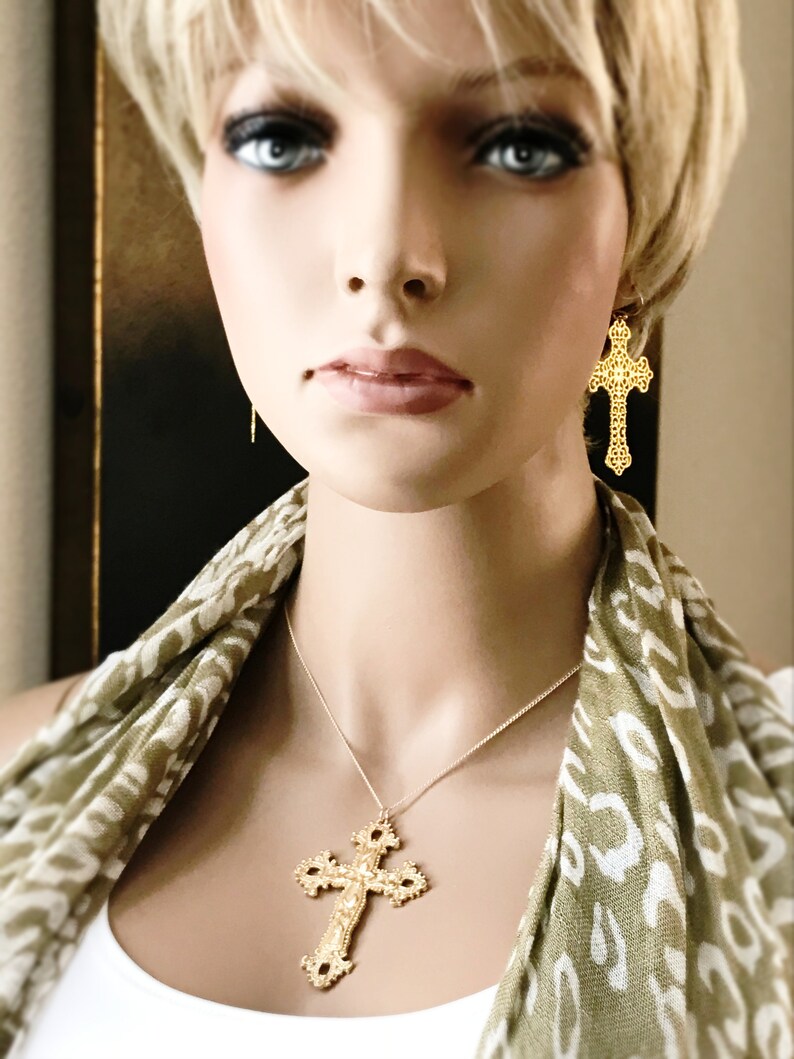 May include: A gold necklace with a large, ornate cross pendant. The necklace is worn by a person with blonde hair and a green and white patterned scarf.