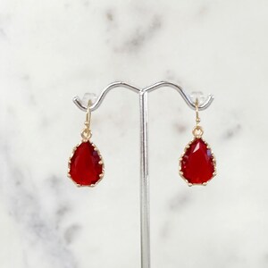May include: A pair of gold-colored drop earrings with teardrop-shaped, faceted red gemstones. The earrings are displayed on a silver jewelry stand against a white background.