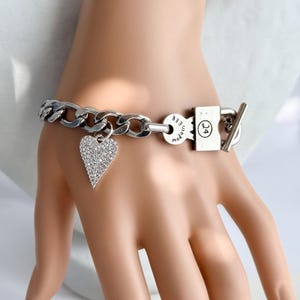 May include: A silver-toned chain link bracelet featuring a heart-shaped charm encrusted with small clear stones. The bracelet includes a rectangular clasp component engraved with the number 24 and circular text reading Made in Italy.