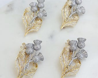 May include: Four thistle-shaped brooches featuring silver-toned flower heads with a grid pattern and gold-toned leaves encrusted with clear crystals. Each brooch displays a detailed botanical design with three distinct thistle heads and foliage.
