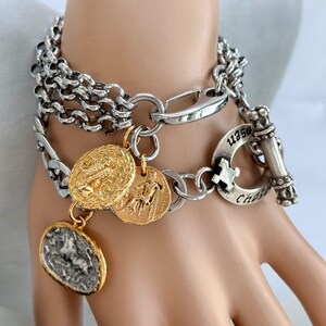 May include: A silver-toned chain link bracelet featuring a toggle clasp with engraved text and three dangling coin-style charms. Two charms are gold-toned with embossed figures, and one charm is a textured silver-toned oval piece.