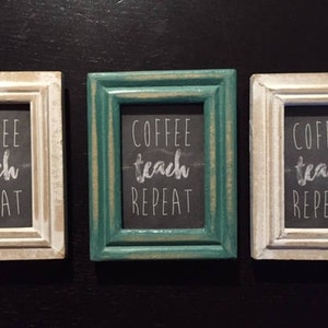 May include: Three white wood framed signs with black chalkboard inserts. Each sign has the text "Coffee Teach Repeat" written in white chalk.