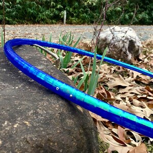 May include: A blue and green hula hoop rests on a dark gray rock. The hoop has a vibrant, translucent design with shades of blue and green. The background features foliage, grass, and fallen leaves, suggesting an outdoor setting.