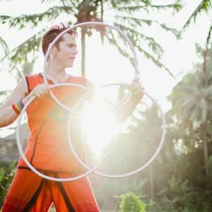 May include: A person in an orange shirt is juggling three silver hoops. The person is outdoors with a bright, sunny background.