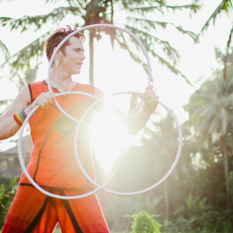 May include: A person in an orange shirt is juggling three silver hoops. The person is outdoors with a bright, sunny background.